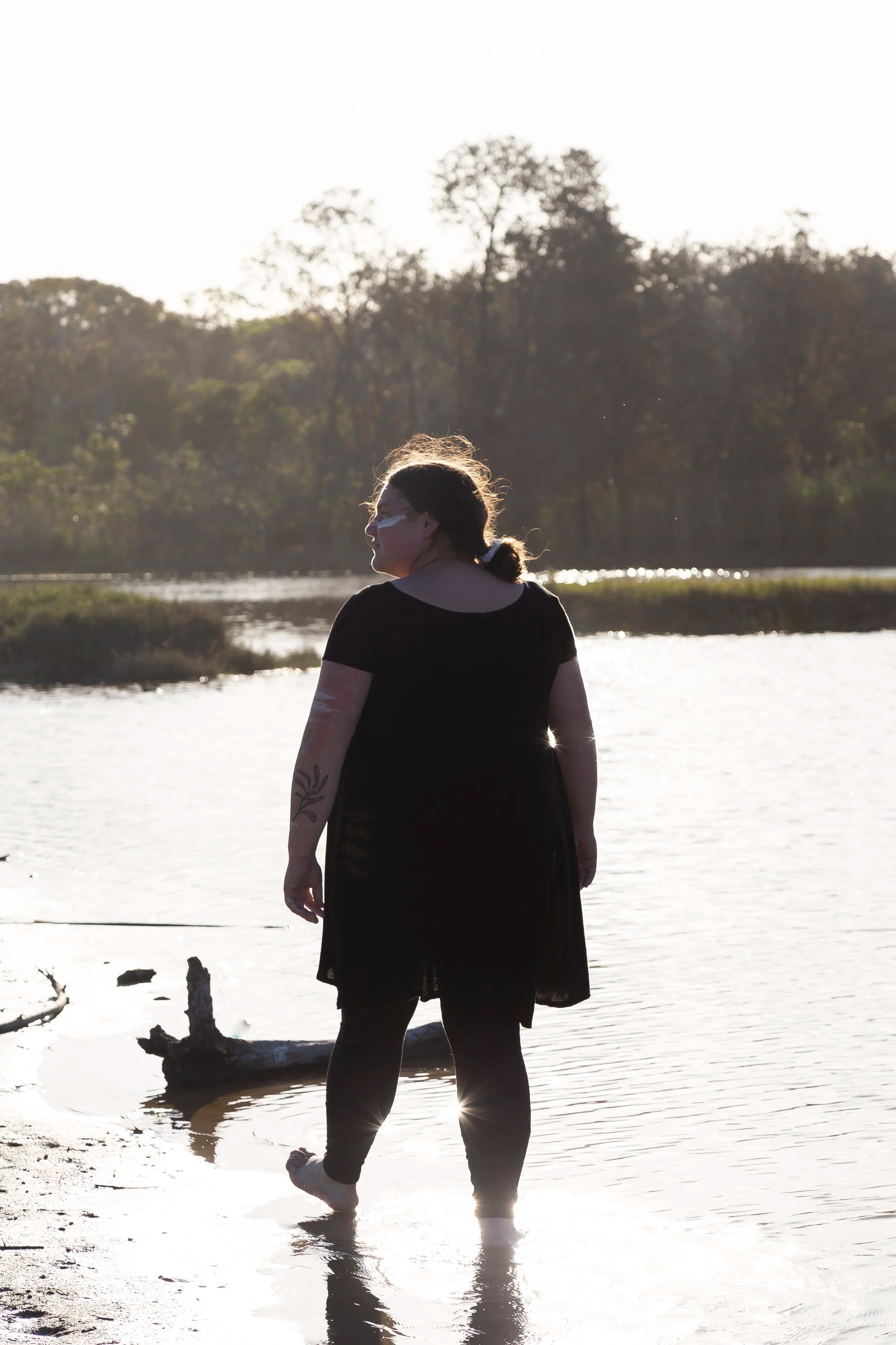 Bundjalung artist, Nickolla Clark, stands barefoot in water at sunset, with ochre markings honouring Arakwal Country and culture.